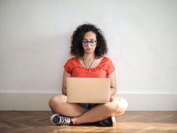 woman in orange crew neck t shirt sitting on brown wooden floor using laptop